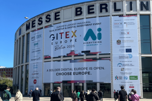 Cilvēki dodas iekšā Berlīnes ēkā, kur notiek Gitex Europe 2025 konference. Uz ēkas redzams konferences plakāts.
