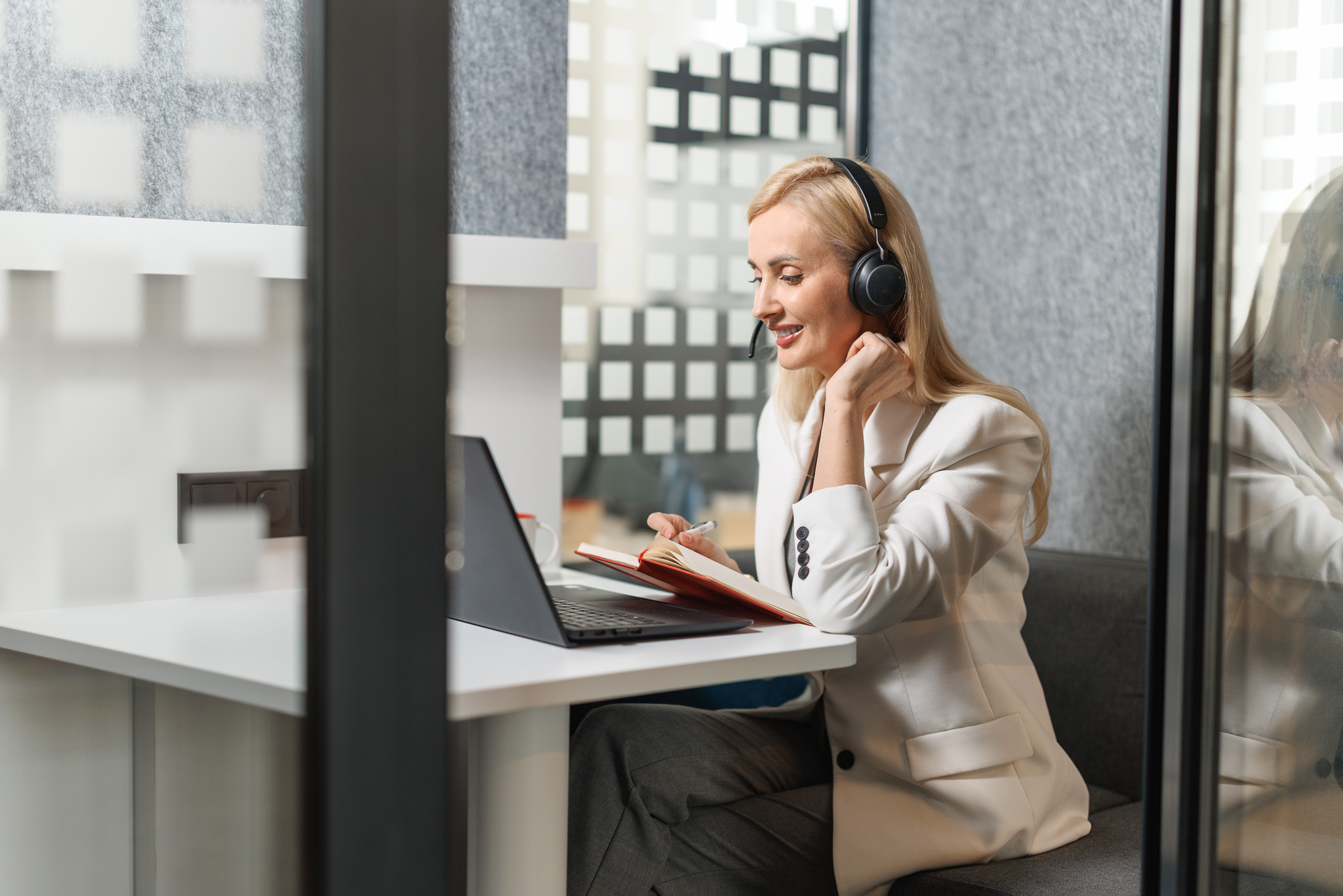 Sieviete ar blondiem matiem un gaišu žaketi un austiņām skatās datorā